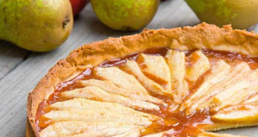 A pie with sliced pears and a glossy glaze is presented on a wooden surface, with fresh pears in the background.