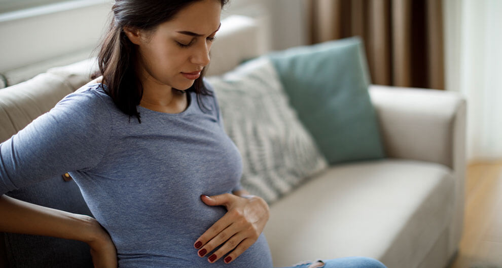 A pregnant woman in a blue shirt sits on a couch, her hand on her belly.
