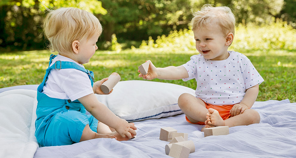 Two toddlers play with wooden blocks on a blanket in a park.