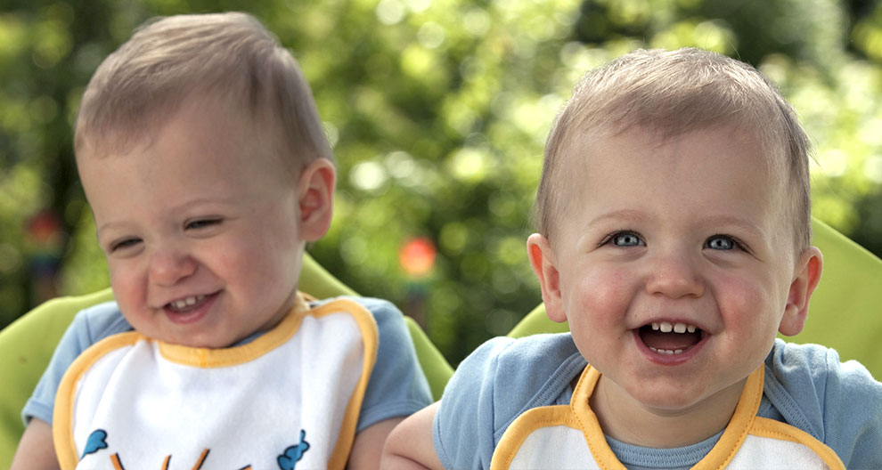 Two toddlers, one with a big smile and the other with a squinted smile, are seated outdoors.